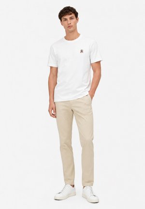 Jeune homme portant un t-shirt blanc à manches courtes, un pantalon beige et des baskets blanches, debout avec une main dans la poche.