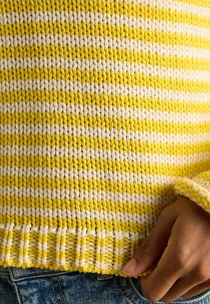 Prim-plan cu o mână sprijinită pe tivul unui pulover tricotat cu dungi galbene și albe, purtat cu blugi din denim albastru.