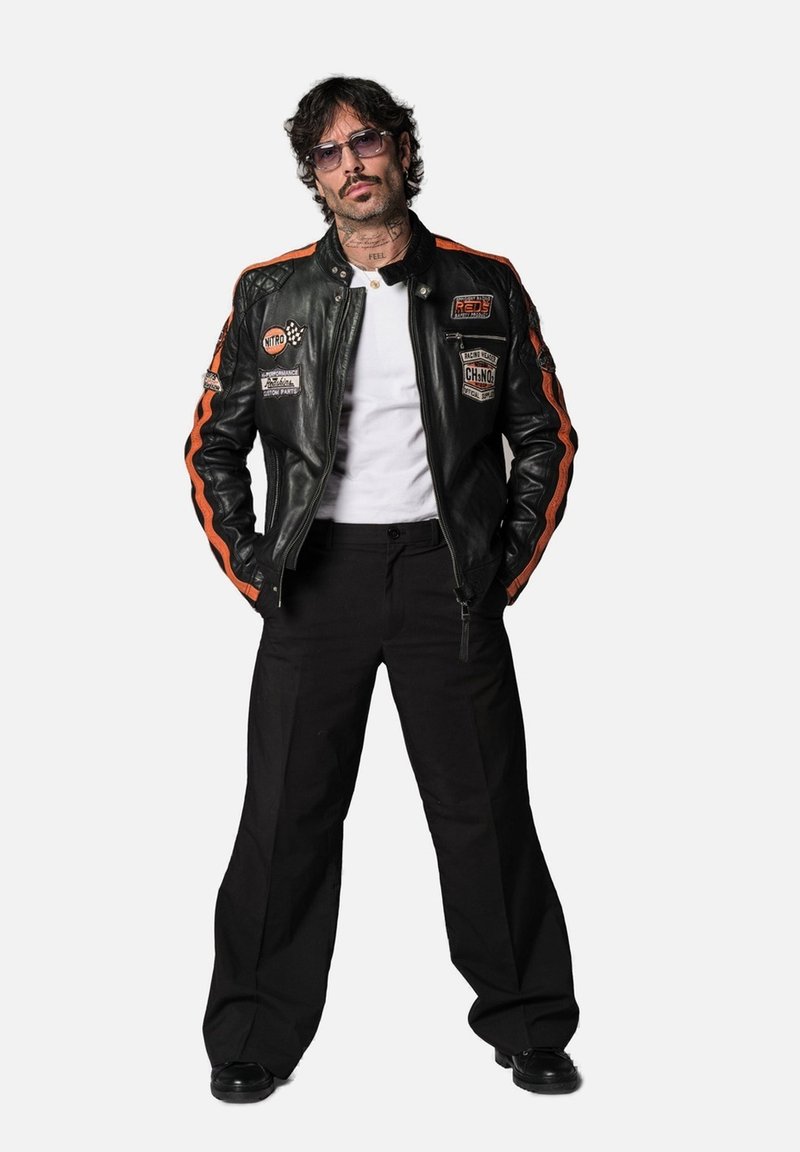 Veste de moto en cuir noir avec des rayures orange, divers écussons, portée sur une chemise blanche. Pantalons larges noirs et chaussures noires.