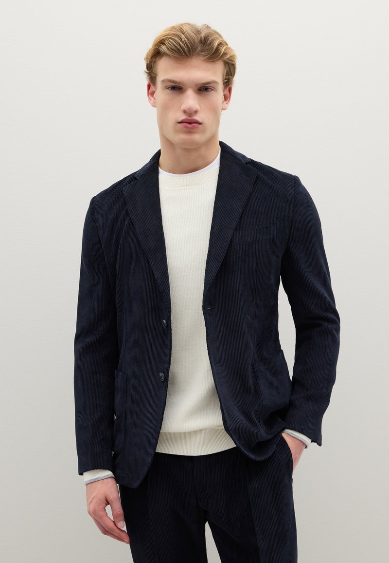 Blazer blu navy in velluto a coste con rever sciallato, due tasche frontali; indossato sopra un maglione leggero a coste con scollo tondo. Tessuto liscio, vestibilità sagomata.