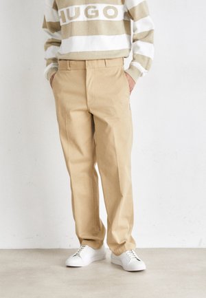 Person, die beige Hose, weiße Sneakers und einen gestreiften beige-weißen Pullover mit dem Schriftzug „HUGO“ auf der Brust trägt.