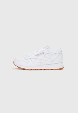 Zapatilla blanca de cuero Reebok con suela de goma, cordones y logo de Reebok con la bandera del Reino Unido en el lateral, mostrada de perfil sobre un fondo liso.