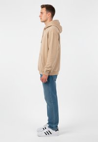 Ung man som står på sidan iförd en beige huvtröja, blå jeans och vita sneakers med svarta ränder mot en helt vit bakgrund.