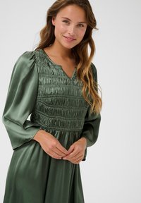 Green satin dress with a smocked bodice, V-neckline, and long sleeves. The fabric has a smooth texture and a flowing design.