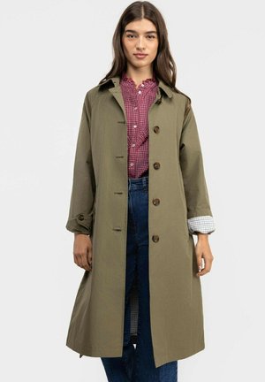 Mujer con un abrigo largo verde oliva abotonado sobre una blusa roja a cuadros y jeans azules, de pie contra un fondo claro y liso.
