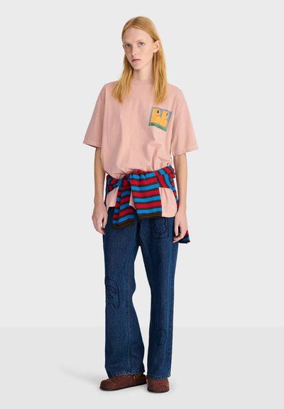 Jeune personne aux longs cheveux blonds porte un t-shirt rose ample, un jean bleu, des chaussures marron et un pull rayé rouge et bleu noué autour de la taille.