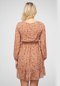 Une robe à manches courtes de couleur rouille avec un motif floral, un taille smockée et un ourlet à volants, fabriquée en tissu léger et semi-transparent.