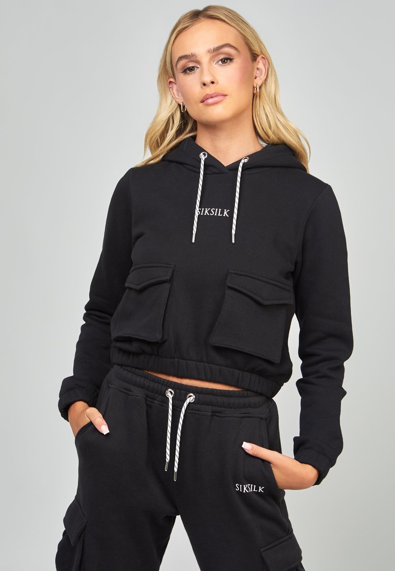 Felpa nera corta in cotone, dotata di due tasche frontali, vita elastica e stringhe argentate. Abbinata a joggers coordinati.