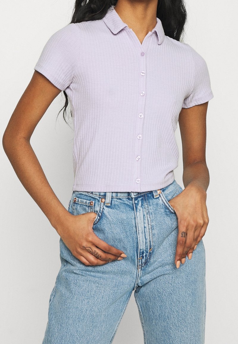 Camisa de manga corta de color lila con textura acanalada y cuello tipo polo, que presenta un cierre frontal de botones y una silueta ajustada, combinada con unos vaqueros de tiro alto de color azul claro.