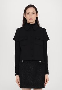 Veste noire raccourcie à manches courtes, avec des boutons sur le devant et deux poches poitrine ; associée à une jupe noire texturée avec des accents métalliques.