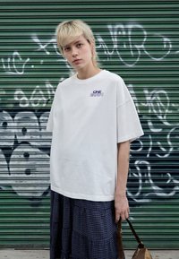 White oversized t-shirt with "ONE EARTH" in purple lettering on the chest, paired with a dark plaid skirt. Background shows green metal shutters.