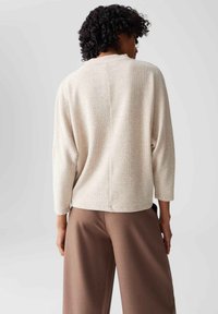 Persoon met krullend haar, gekleed in een gestructureerd beige lange mouw top en losse bruine broek, met de rug naar de camera tegen een effen lichte achtergrond.