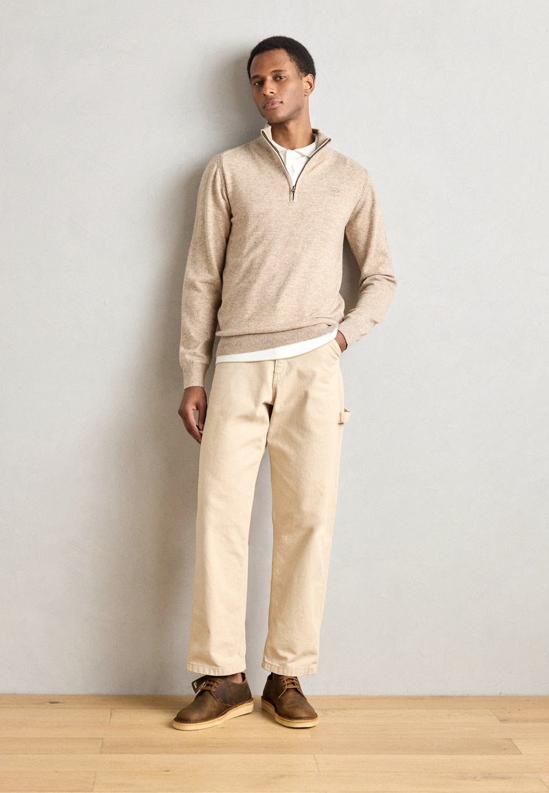 Suéter de algodón color beige con cremallera, sobre una camisa blanca, combinado con pantalones beige y zapatos de cuero marrón. Diseño simple y paleta de colores neutros.
