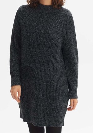 Kvinde iført en mørkegrå, langærmet, ribstrikket sweaterkjole, stående mod en ensfarvet hvid baggrund.