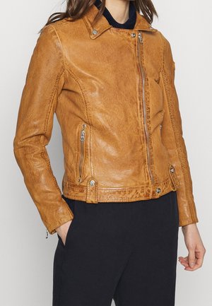 Frau trägt eine braune Bikerjacke aus Leder mit Reißverschlusstaschen und asymmetrischem Frontreißverschluss, kombiniert mit schwarzer Hose.