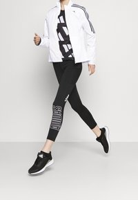 Chaqueta de chándal blanca con rayas negras, camiseta gráfica negra, leggings negros con el texto "ADIDAS" y zapatillas deportivas negras con suelas blancas.