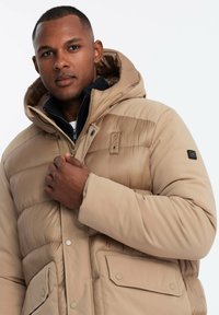 Beige Pufferjacke mit Kapuze, ausgestattet mit einem Reißverschluss, Druckknöpfen und Fronttaschen. Glatte, leicht glänzende Textur mit Steppmuster.