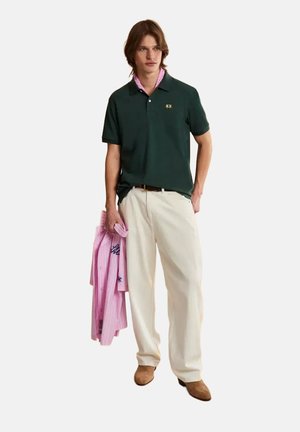 Young man in dark green polo, cream trousers, brown shoes, holding pink striped shirt with embroidery, standing against plain background.