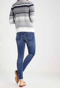 Kvinna klädd i blåa skinny jeans, beiga platta skor och en mönstrad stickad tröja med fransar vid fållen, stående med ett ben korsat.