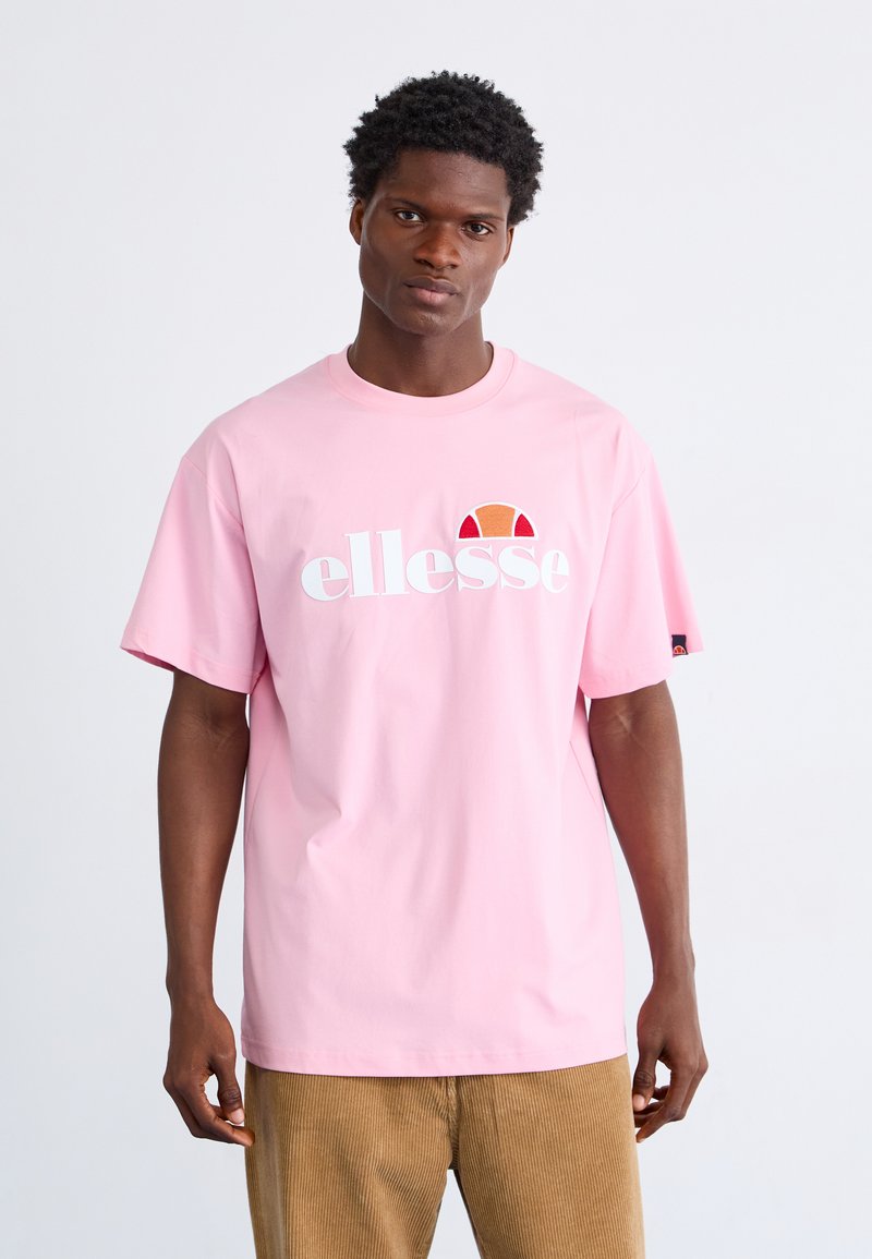 Rozā kokvilnas T-kreklā ar brīvu piegriezumu, kas ir ar baltu "ellesse" logo un krāsainu pusapļa grafiku virs teksta. Īsās piedurknes.