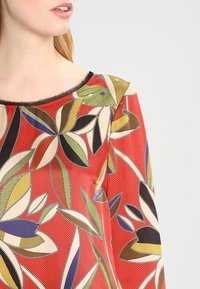 Flerfärgad blommig topp i röd med gröna, gula och blå blad. Har rund halsringning och textil med struktur samt svarta detaljer.