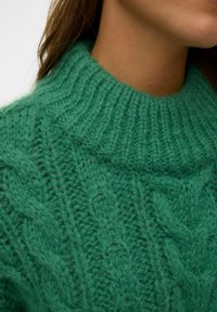 Nahaufnahme einer Person, die einen grünen, grob gestrickten Pullover mit einem gerippten hohen Kragen trägt.