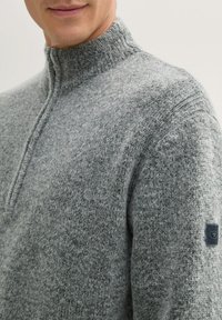 Grau gestrickter Pullover mit hohem Kragen, strukturiertem Stoff und einem kleinen Logopatch am linken Ärmel. Weiches, warmes Aussehen.