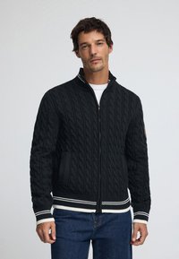 Tungstickad zip-up-kofta i mörkgrått med kabelmönster, ribbad fåll och muddar med vita och svarta ränder samt sidofickor.