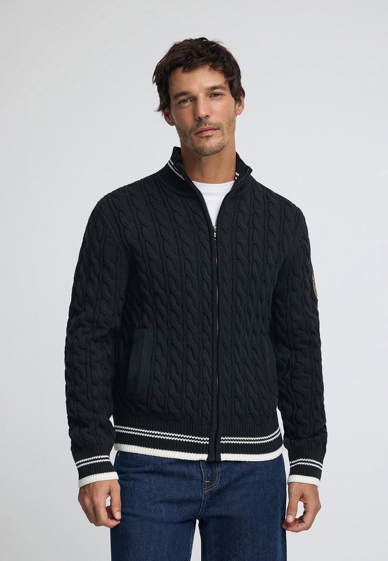 Tungstickad zip-up-kofta i mörkgrått med kabelmönster, ribbad fåll och muddar med vita och svarta ränder samt sidofickor.