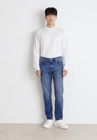 White long-sleeve shirt paired with blue jeans. Model wears white sneakers. Simple design with a relaxed fit and minimal detailing.