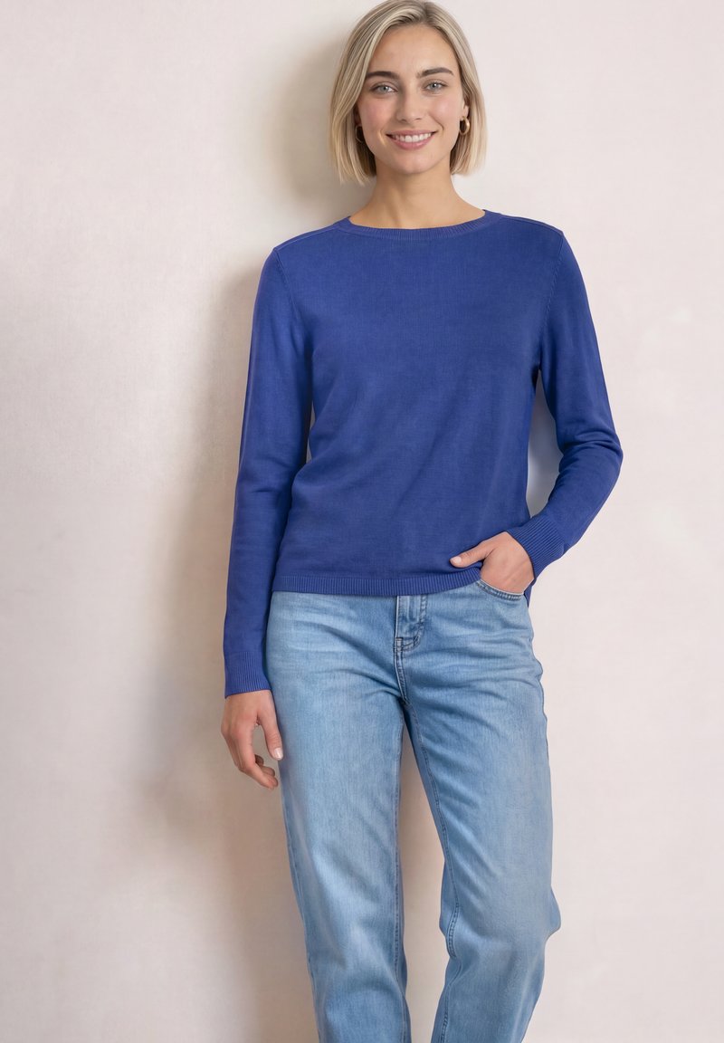 Jeune femme aux cheveux blonds courts portant une chemise bleue à manches longues et un jean bleu clair, souriante avec une main dans la poche.