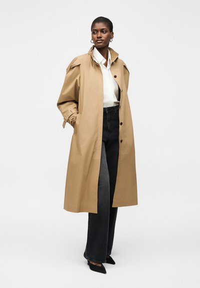 Femme portant un long trench beige sur une chemise blanche et un jean noir, debout les mains dans les poches du manteau devant un fond uni.