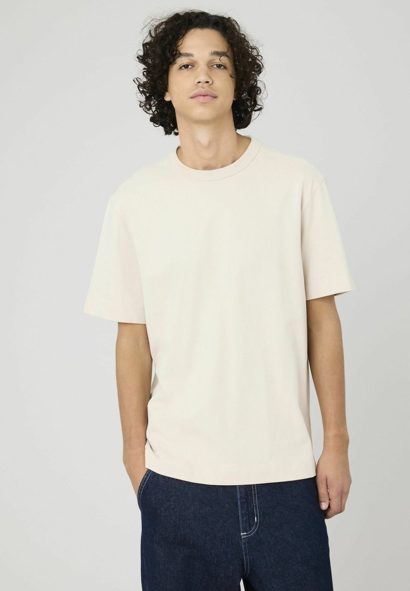 T-shirt en coton beige à manches courtes, col rond et coupe décontractée. Associé à un jean bleu, avec une coupe droite et des coutures contrastées.
