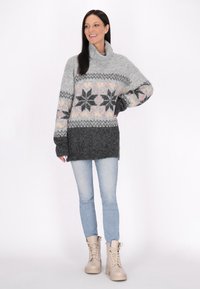 Oversized gebreide trui in grijs met roze, zwart en crème patronen. Heeft een funnelnek en lange mouwen. Gecombineerd met lichtblauwe jeans en beige laarzen.