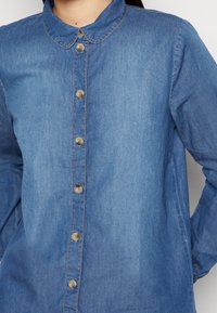 Camicia di denim azzurra con colletto arrotondato, patta con bottoni frontale e maniche lunghe. Presenta una consistenza morbida e dettagli di cucitura delicati.