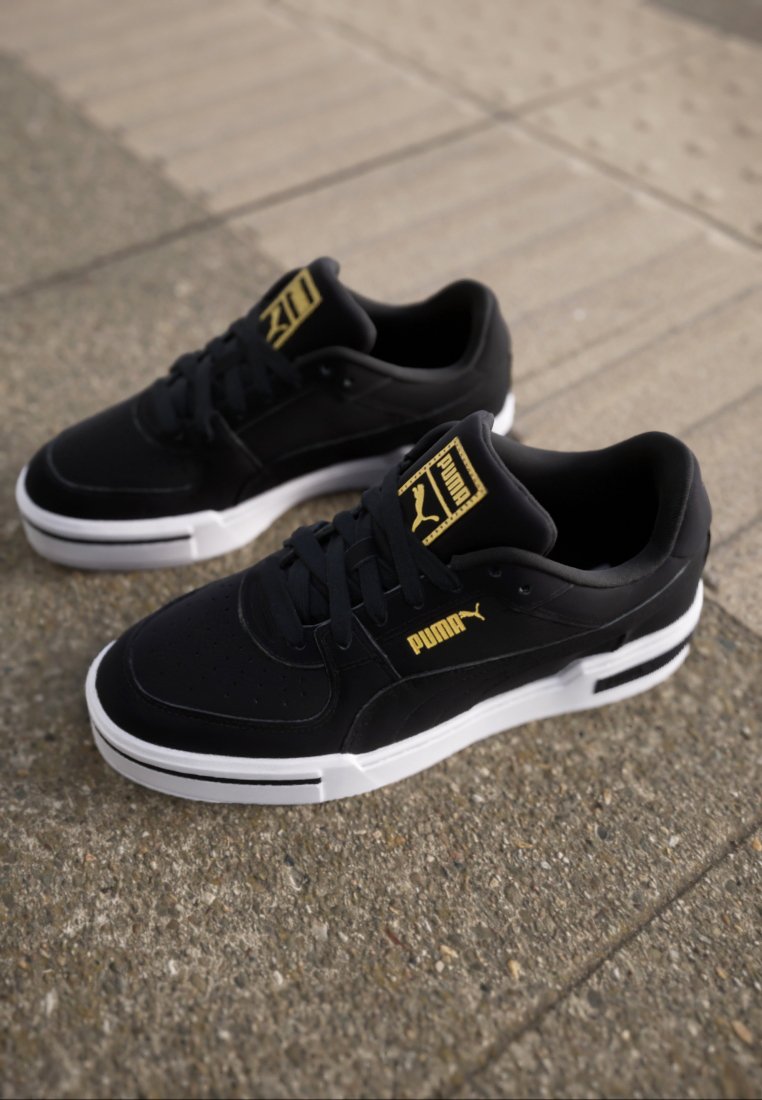 Baskets noires avec une tige en cuir lisse, un col rembourré, une semelle en caoutchouc blanche et des accents dorés présentant le logo et le branding Puma.