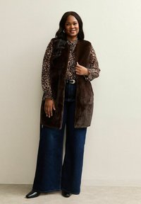 Brązowa kamizelka z sztucznego futra z otwartym przodem na wzorzystą bluzkę. Szerokie, ciemnoniebieskie dżinsy i czarne buty na szpilkach. Gładka tekstura, casualowy styl.