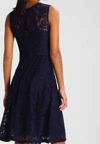 Navy blue lace dress with sleeveless design, textured floral patterns, and a back zipper closure. A-line silhouette with a knee-length hem.
