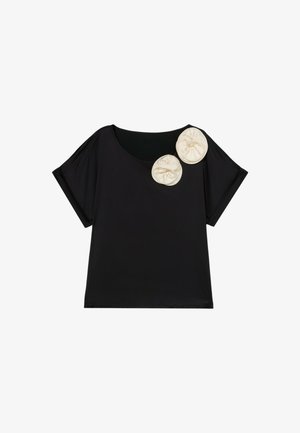 Blusa negra de manga corta con escote redondeado y dos grandes adornos de tela color crema en el hombro derecho.