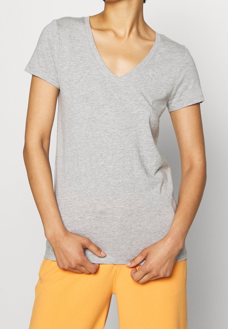 Personne portant un t-shirt gris clair à col en V et manches courtes, et un pantalon jaune moutarde, les mains saisissant délicatement l'ourlet du t-shirt.