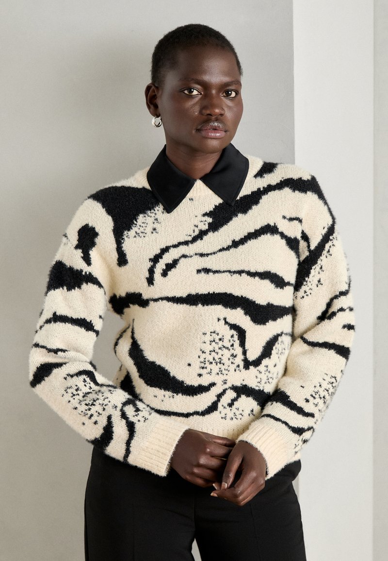 Strickpullover in Creme mit schwarzem abstraktem Muster, mit einem schwarzen Kragen, langen Ärmeln und gerippten Bündchen.