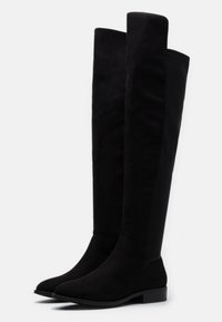 Botas negras por encima de la rodilla con una suave textura de ante, forma esbelta, tacón plano y un diseño de panel lateral en contraste con un acabado mate.