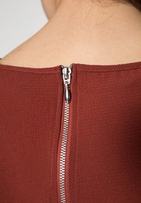 Gros plan d'une fermeture éclair argentée sur le dos d'un vêtement texturé rouge foncé porté sur une peau nue.