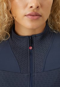 Veste en polaire marine avec un col haut, présentant un devant texturé, une fermeture éclair et un accent en métal rouge près de la tirette de la fermeture.