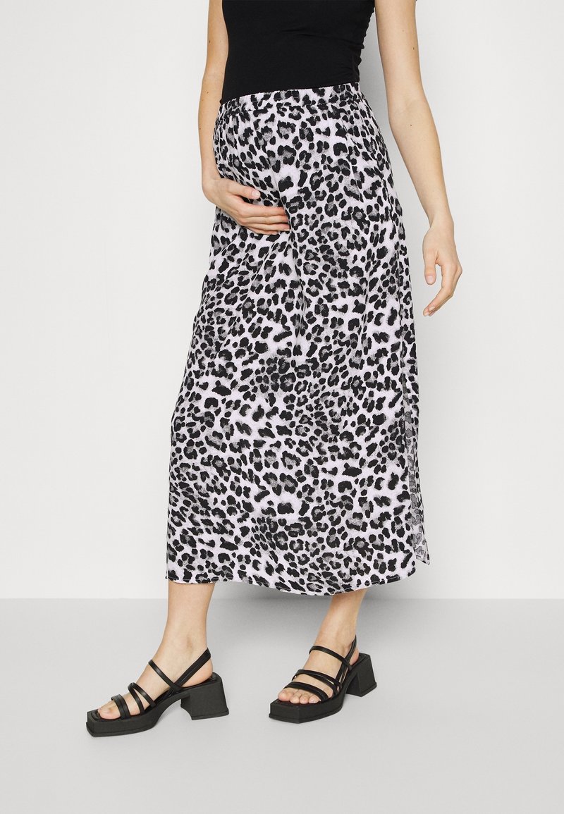 Femme portant un haut noir, une jupe midi imprimé léopard noir et blanc, et des sandales noires à talons blocs avec des brides, debout sur un sol gris.