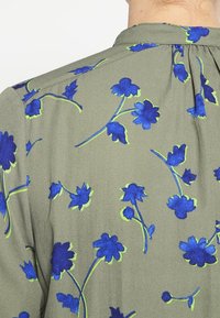 Olivově zelená blůza se květinovým vzorem v modré s zelenými akcenty, hedvábná textura, řasený zadní detail a klasický límec.