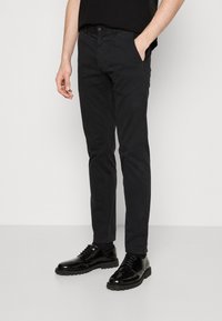 Black, slim-fit trousers with a smooth texture, featuring side pockets and a button closure, paired with glossy black lace-up shoes.