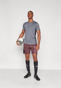 Grått åtsittande atletisk tröja med mörka sidopaneler, vinröda shorts med geometriskt mönster, svarta strumpor och svarta fotbollsskor, håller i en fotboll.
