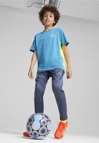 Sininen ja keltainen urheilupaita tekstuurilla, harmaat housut ja kirkkaan oranssit jalkapallokengät. Seisoo kuvioidun jalkapallon päällä.