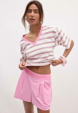 Frau trägt ein weiß-rosa gestreiftes V-Ausschnitt-Shirt, leicht angehoben, und hoch taillierte rosa Shorts, accessorisiert mit klobigen beigen und rosa Armbändern.